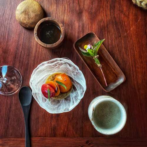 A wooden table with a dish of colorful fruit, a small flower, a cup of coffee, and a glass of water.
