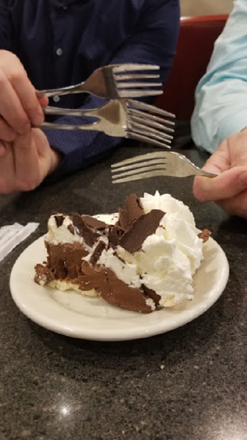 Two hands with forks reaching for a slice of chocolate dessert topped with whipped cream on a plate.
