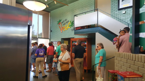 A busy indoor space with people waiting in line, colorful decor, and a staircase in the background.