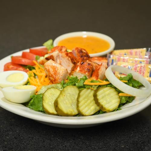 A colorful salad plate with grilled chicken, tomatoes, pickles, hard-boiled eggs, cheese, and a side of dressing.