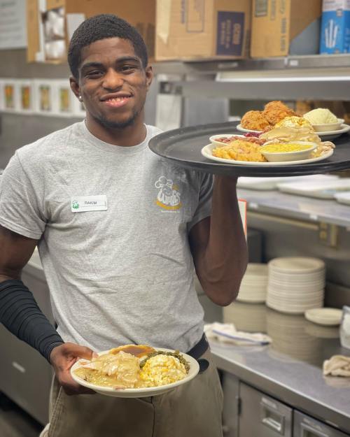 A young man in a gray shirt smiles while holding two trays of food in a busy kitchen.