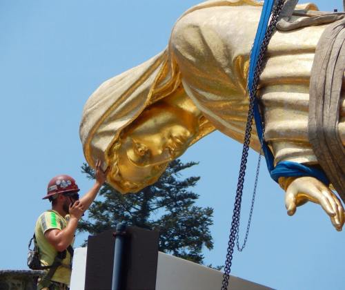 A worker adjusts a large golden statue of a woman, with trees and blue sky in the background.