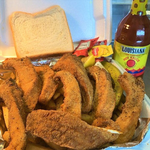A takeout container filled with fried fish, bread, pickles, and hot sauce bottles on the side.