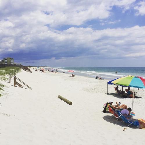 A sunny beach scene with people relaxing under umbrellas, a colorful tent, and gentle waves under a cloudy sky.