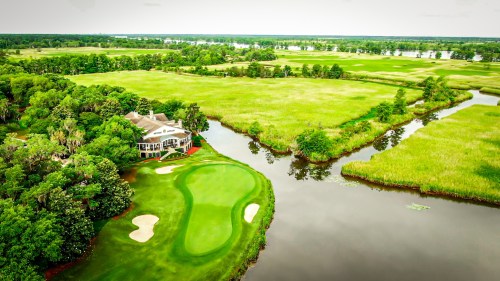 Aerial view of a golf course with a clubhouse, surrounded by lush greenery and a winding river.