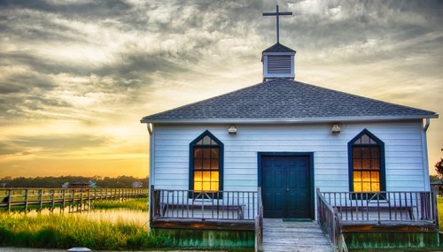 A quaint white church with a cross on top, surrounded by marshland and a colorful sunset sky.