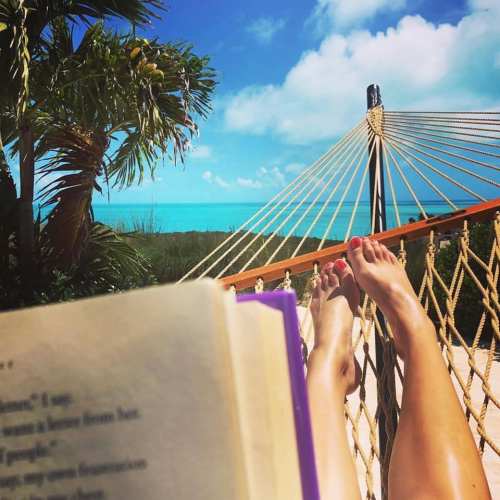 A person relaxes in a hammock by the beach, reading a book with turquoise water and palm trees in the background.