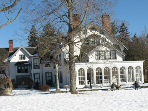 A large white mansion with multiple gables, surrounded by snow and trees, featuring a sunroom and people sitting outside.
