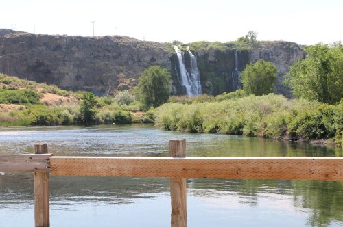 A serene river scene with a wooden railing in the foreground and a waterfall cascading down rocky cliffs in the background.
