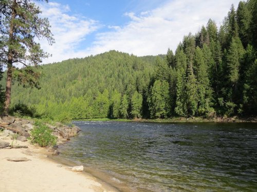 A serene river flows beside a sandy shore, surrounded by lush green trees and a mountainous backdrop under a blue sky.
