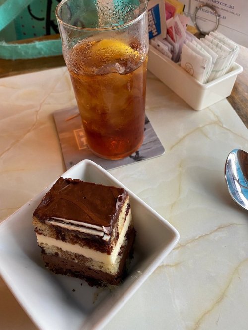 A slice of layered chocolate cake on a small plate next to a glass of iced tea on a table.