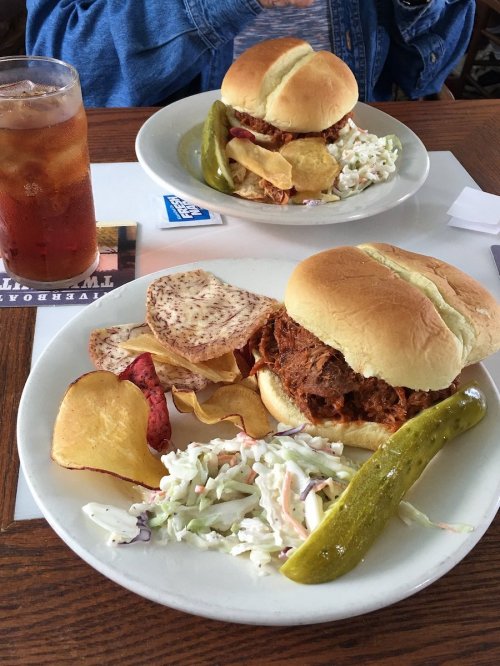 Two pulled pork sandwiches on plates, served with coleslaw, chips, and a pickle, alongside a glass of iced tea.