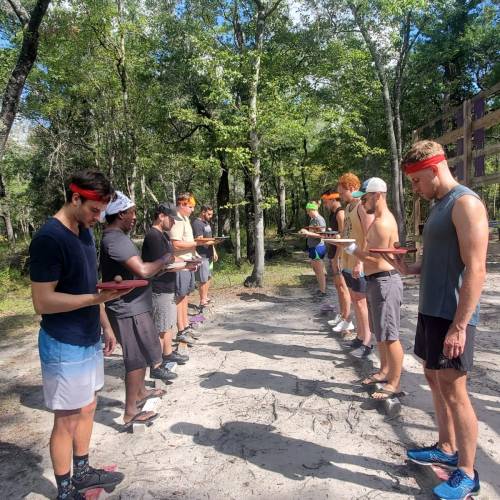 A group of men in athletic wear stand in two lines outdoors, holding plates, surrounded by trees and sunlight.