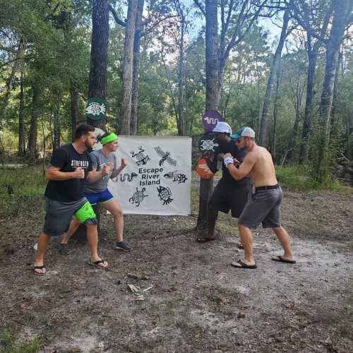 Four people playfully pose in a forest, facing each other with a sign that reads "Escape River Island" in the background.