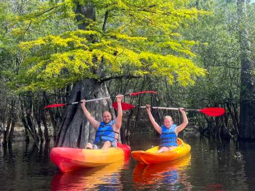 Two people in colorful kayaks raise their paddles in celebration on a calm, tree-lined waterway.