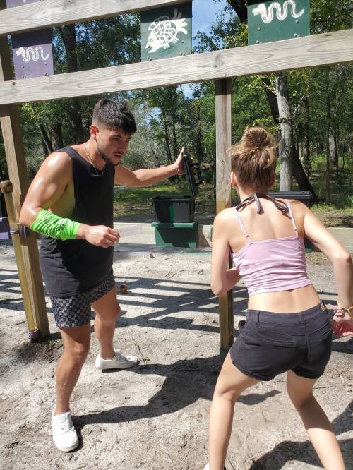 Two young adults engage in a playful activity at a park, with a wooden structure in the background.