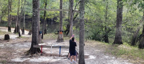 A person swings on a wooden structure in a wooded area near a calm body of water. Trees surround the scene.