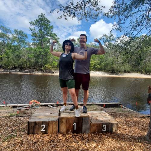 Two people flexing their muscles on a podium by a river, surrounded by trees and a clear blue sky.