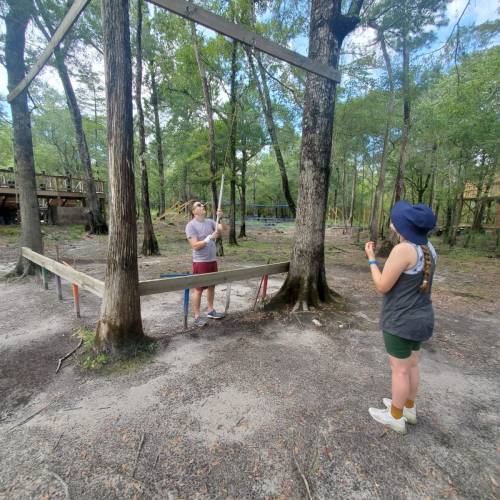Two people in a wooded area, one adjusting a wooden structure while the other watches, surrounded by trees.