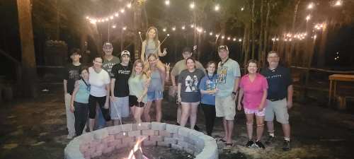 A group of people gathered around a campfire at night, with string lights illuminating the trees in the background.