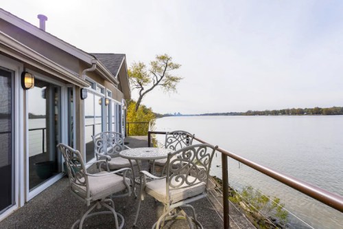 A serene lakeside patio with a round table and chairs, overlooking calm waters and distant city skyline.