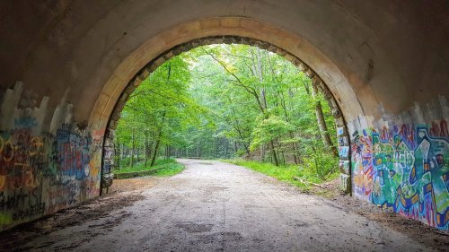 A view through a stone archway into a lush, green forest with a dirt path and colorful graffiti on the walls.