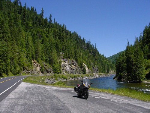 A motorcycle parked by a scenic road alongside a river, surrounded by lush green trees and mountains under a clear blue sky.