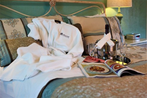 A cozy hotel bed with a robe, champagne, glasses, and a magazine on top, creating a relaxing atmosphere.