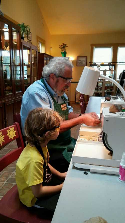 An older man and a young boy work together at a table, focused on a craft project in a bright, sunny room.