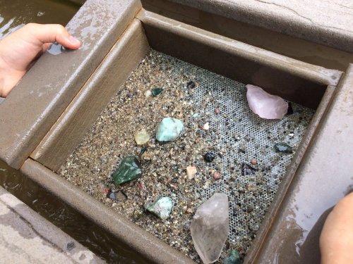 A person holds a tray filled with sand and various colorful gemstones, partially submerged in water.
