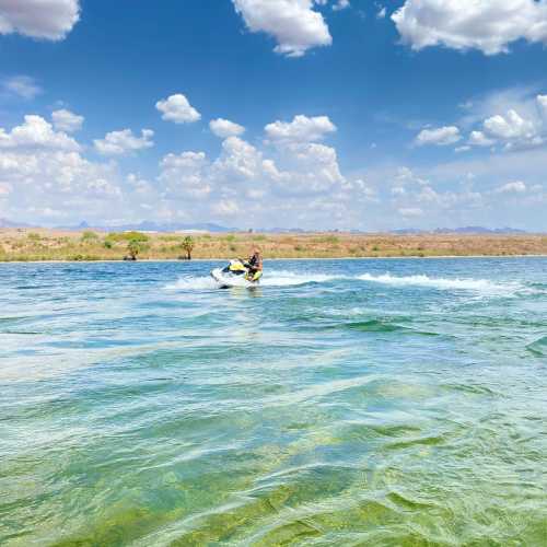 A person rides a jet ski on a clear blue river under a bright sky with fluffy clouds and distant hills.