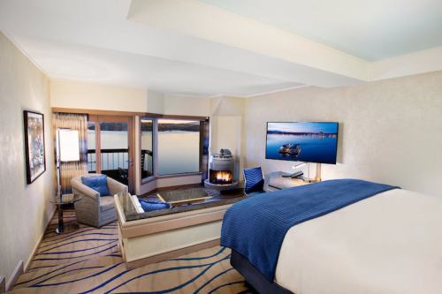 Cozy hotel room with a lake view, featuring a bed, seating area, fireplace, and large TV.