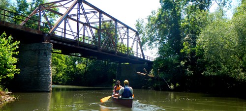 Two people canoeing under a rustic bridge surrounded by lush greenery and calm waters.