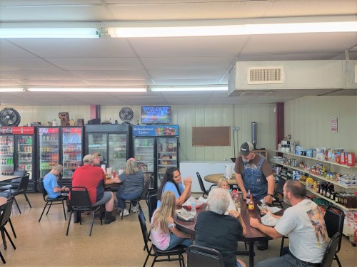A casual dining area with people eating at tables, surrounded by shelves of snacks and drinks. A TV is visible in the background.
