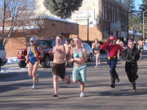 A group of people in swimwear running down a snowy street, with some in costumes and a few cars parked nearby.