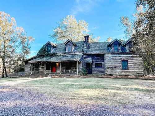 A rustic, weathered wooden house with a porch, surrounded by trees and a clear blue sky.