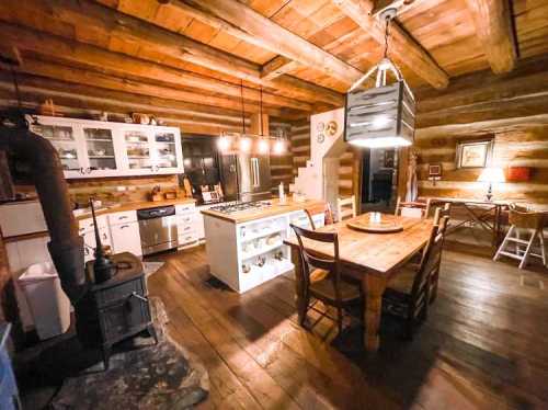 Cozy log cabin kitchen with wooden beams, a dining table, modern appliances, and warm lighting.