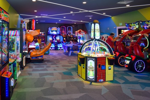 A vibrant arcade filled with colorful games, including racing machines and claw machines, under bright lights.