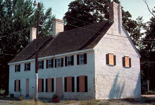 Abandoned two-story house with boarded windows, a sloped roof, and multiple chimneys, surrounded by trees.