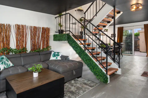 Modern living room with a gray sectional sofa, wooden stairs, and decorative plants along the walls and staircase.