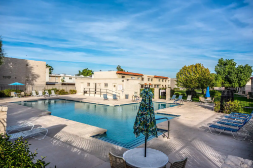 A sunny pool area with lounge chairs, umbrellas, and surrounding greenery in a residential complex.