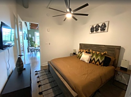 Cozy bedroom with a wooden bed, decorative pillows, a TV, and a ceiling fan, leading to a bright living area.