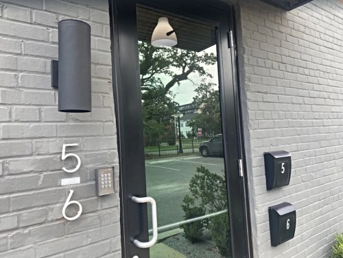 A modern entrance door with a black handle, intercom, and mailboxes numbered 5 and 6, set against a gray brick wall.