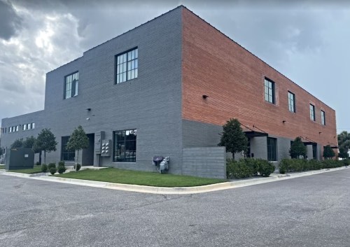 A modern two-story building with a mix of gray and red brick, surrounded by greenery and a paved parking area.
