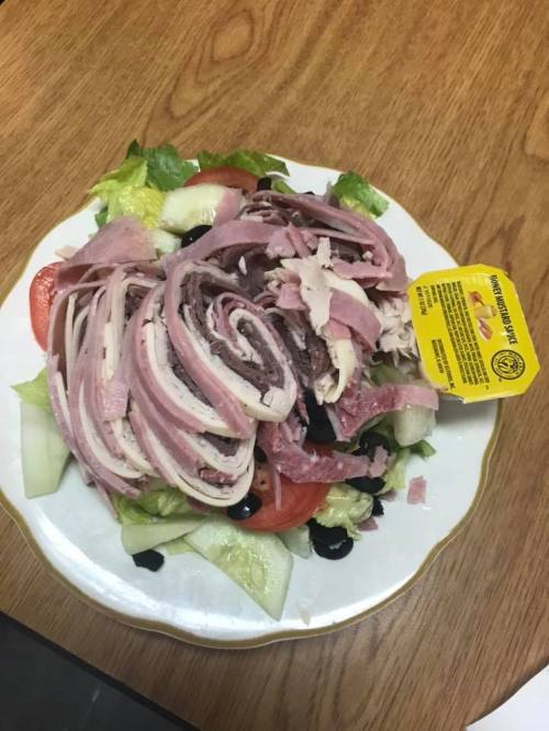 A plate of salad topped with rolled deli meats, tomatoes, cucumbers, and a small container of dressing.