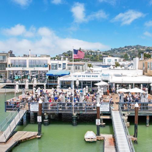A bustling pier with diners and an American flag, surrounded by water and buildings under a clear blue sky.