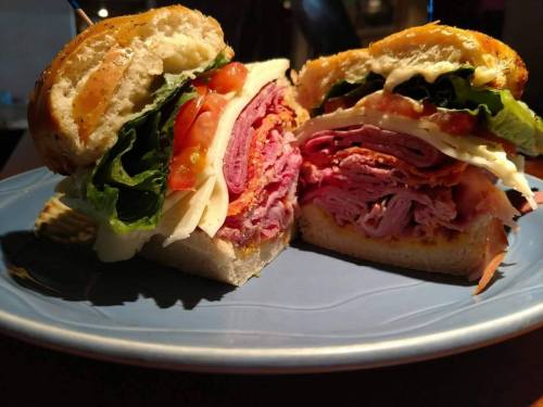 A close-up of a sandwich cut in half, featuring layers of meats, lettuce, tomato, and cheese on a toasted bun.