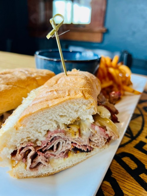 A close-up of a sandwich filled with meats and cheese, served with sweet potato fries and a small bowl of sauce.