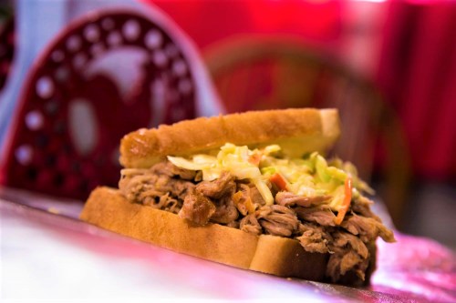 A close-up of a pulled pork sandwich with coleslaw on two slices of bread, set against a blurred background.