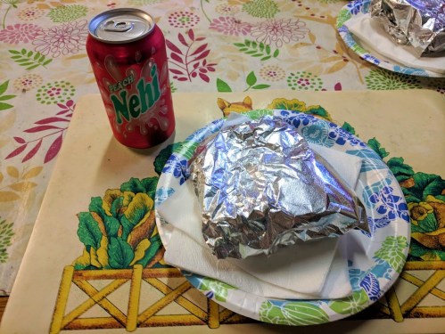A can of Peach Nehi soda next to a foil-wrapped sandwich on a paper plate, set on a floral tablecloth.